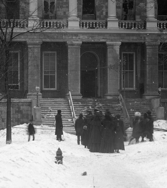 View of the crowd standing at the South Wing of the Wisconsin State Capitol watching the fire that destroyed most of the building. Because the University well on which the Capitol depended for water had been drained shortly before the fire, the useless fire hydrant in the foreground is an ironic element.