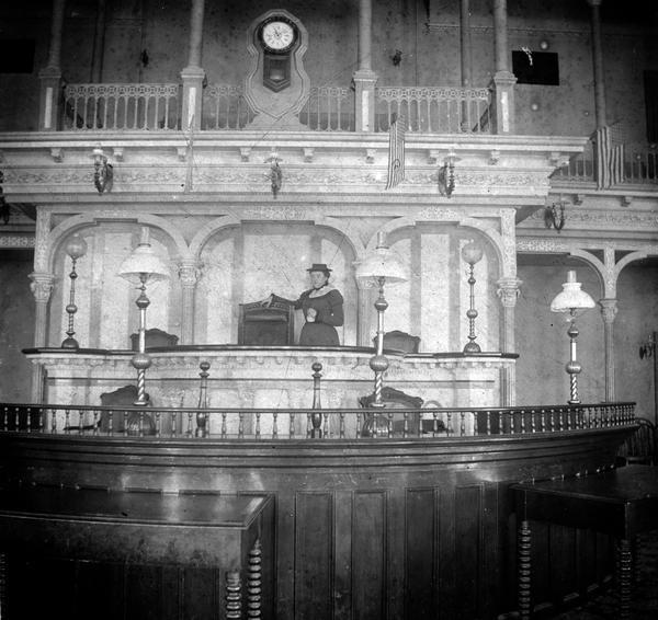 Assembly Speaker's desk, showing an unidentified woman, probably a tourist, standing by the Speaker's chair. In front of the Speaker's desk are the clerks' desk and the journalists' tables. This photograph, the original of which is a small faded snapshot that has been digitally restored, is the only known photograph to document this part of the Assembly Chamber. The Speaker's chair is the same as the one in the 1899 photograph, but the Eastlake chairs in this photograph had recently been replaced. These chairs match those in the Supreme Court Chamber (Image ID: 3477) and the Board of Control Office (Image ID: 10504), both located in the 1883 additions, and they probably represent furnishing purchased at that time.