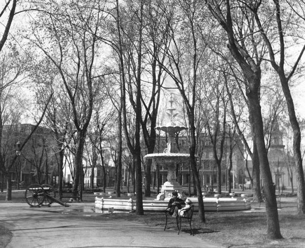The replica of the Centennial Fountain in the Capitol Park, where people relax on a park bench. This view looks toward the intersection of Main and Carroll streets, with the Park Hotel and the First Baptist Church visible through the trees.