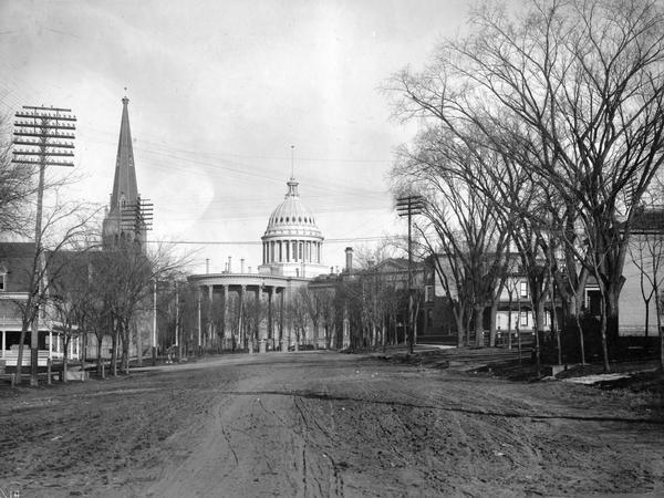 The third Wisconsin State Capitol as seen from unpaved West Washington Avenue, with the West Wing to the left and the South Wing to the right. The church spire to the left is Grace Episcopal Church, one of two buildings dating from the 1850s still standing on the Capitol Square.