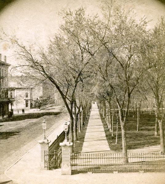Slightly elevated view of the stone gateway at the intersection of Pinckney, Main, and King Streets and the iron fence that surrounded the entire Capitol Park area from 1873 until 1899. This image illustrates the more formal and dignified appearance desired for the park during the later Victorian era. The Vilas House hotel is almost out of the picture on the left. This photograph was distributed by Photographer Isaacs as part of his Lake City Views series.