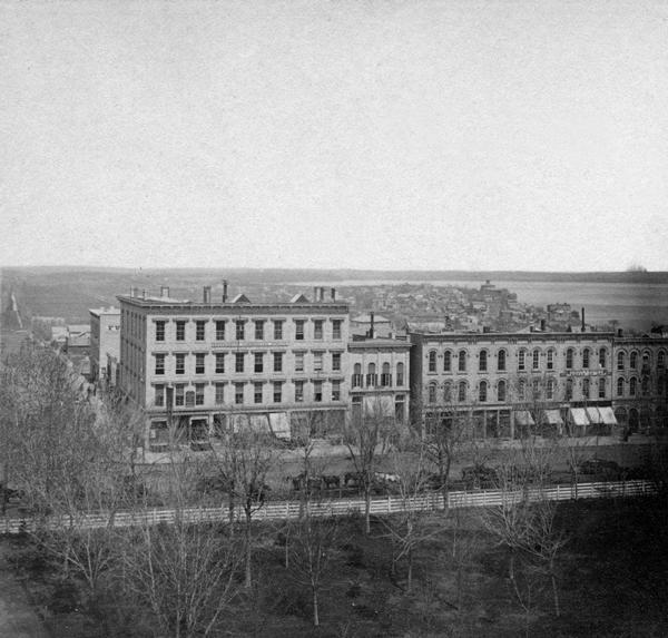 Elevated view of South Pinckney Street from the Wisconsin State Capitol, with East Washington Avenue to the left and Lake Monona on the distant right. The Capitol Park in the foreground is surrounded by the wooden fence that was replaced with an iron fence in 1873, and horse and wagon teams are hitched to the rail that ran around the outside of the wooden fence.