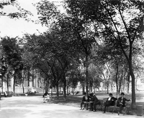 An idyllic afternoon in the Capitol Park, showing groups of men and women seated on benches and conversing. The reproduction Centennial fountain is almost out of view at the left.
