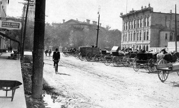 View of the North Wing of the third Wisconsin State Capitol in the background, the Capitol Park, and farm wagons on East Washington Avenue on market day. To the right is the American Exchange Bank, then the First National Bank, one of two 1850s buildings still standing on the Capitol Square.