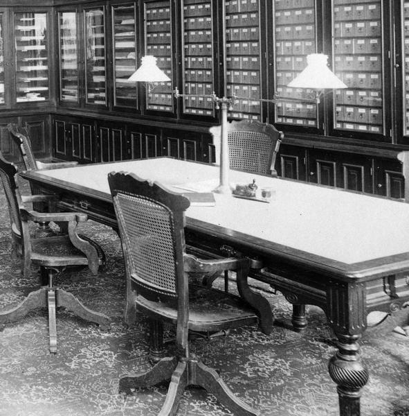 Office of the State Board of Control of Wisconsin Reformatory, Charitable and Penal Institutions, in the third Wisconsin State Capitol, showing details of the Eastlake table, chairs, and gas light fixture, the patterned carpet, and the cabinets for record storage. This office was located in the Capitol addition completed in 1883, and furniture was likely purchased at that time. Identical and similar furniture can be found in other photographs, suggesting that the Eastlake style was prevalent in the additions built in 1883.