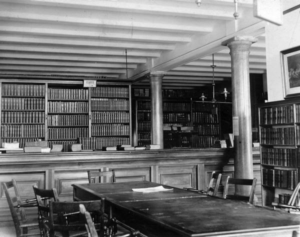 The circulation desk and study area in the Historical Society's library in the third Wisconsin State Capitol. This photograph taken by periodical librarian Mary N. Foster documents the large space in the new South Wing into which the Society moved in 1883. Because the Society's rooms included two levels of library stacks that were closed to the public, the ceiling between the two floors was only roughly finished, a fact which reveals more about the construction of the addition.