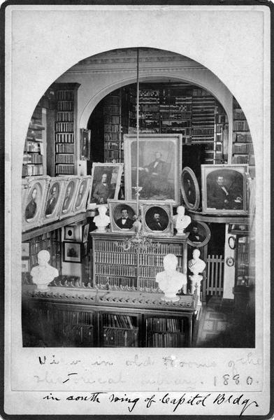 The Historical Society's early library and painting collections on display in the third Wisconsin State Capitol. This photograph was taken about 1880 before the Society moved from the old South Wing (into which it had moved in 1866) to the South Wing addition, and it documents something of the appearance of the original Capitol construction, including the decorative crown moldings, the gas chandelier, and the library cabinets. Carpets are known to have been changed in the Capitol, and it is unlikely that the patterned design here is original.