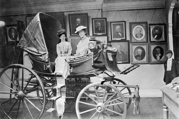 The Daniel Webster carriage on display in the Historical Society's museum in the third Wisconsin State Capitol. At that time the Historical Society's quarters were located in the addition constructed in 1883. Architectural details of the addition that can be gleaned here include the archway to the right and the crown molding from which the Society's painting collection is suspended. The Webster carriage continued to be a favorite exhibit with visitors after the Society moved to its new building on State Street in 1900. Currently, the carriage Webster purchased in 1810 is housed at the Stonefield Village Historic Site in Cassville. The museum visitors are unidentified.