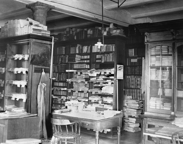 Cluttered work space of the librarian responsible for the Historical Society's newspaper collection during the period when the Society occupied rooms in the third Wisconsin State Capitol. Details of the decoration include the light fixture, the Eastlake desk, and one of the ubiquitous captains chairs. Although the original gas light fixture had been retrofitted for electricity by the time this picture was taken, clearly there was a time when an open gas flame was dangerously close to large quantities of historical paper!