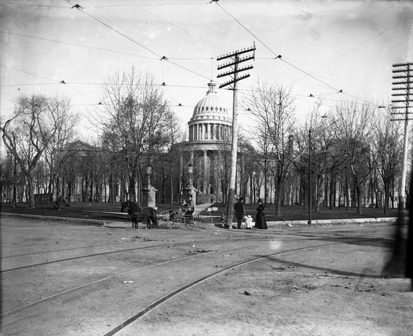 The State Street approach to the Wisconsin State Capitol at the turn of the century. Electric poles and street railroad wires are in the foreground. In the foreground also are members of the William Barckhan family in a carriage pulled a pair of miniature horses, together with an unidentified family group.