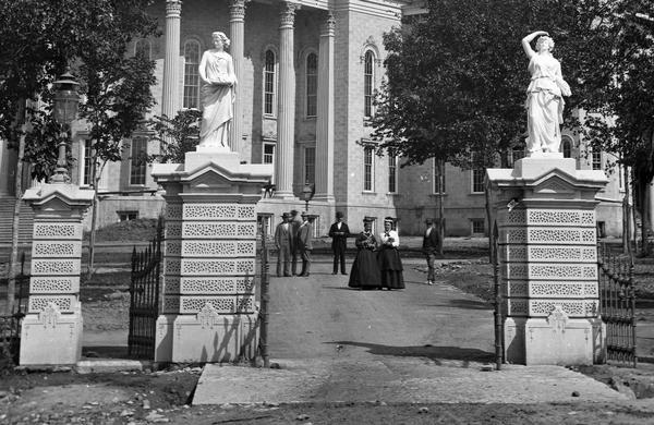 View from street through gates towards a small group in front of the third Wisconsin State Capitol near the West Washington Avenue entrance. The iron fence and stone gateways around the perimeter of the Capitol Park were designed by Stephen V. Shipman, who had also designed the dome, completed in 1873. There are iron sculptures on top of the columns.