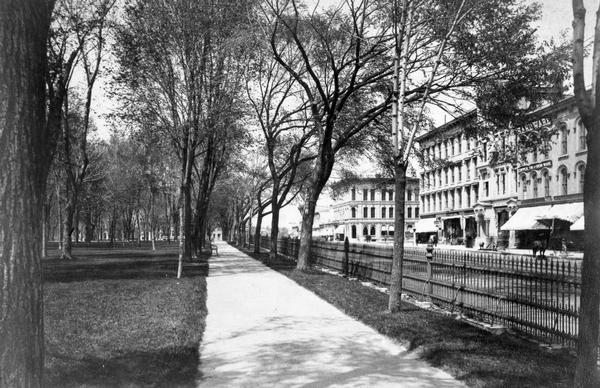 A view of the Capitol Park and Pinckney Street taken approximately 1885-1890. The building in the center, at the intersection of Pinckney and East Washington Avenue, was the First National Bank, now the American Exchange Bank. This pastoral view of the park shows the construction method used to support the iron fence, a less expensive version of the stone coping and foundation proposed in 1872 by Architect Stephen V. Shipman. The iron fence was removed in 1899.