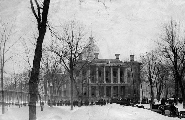 Spectators standing at the South Wing during the aftermath of the fire which destroyed the second Wisconsin State Capitol in Madison, February 26-27, 1904. Although the fire appears to be out in this photograph, smoke still obscures the dome. The pathways are littered with tables and chairs and books that have been rescued. 
