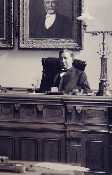 Detail of the 1903 group portrait of the Wisconsin Supreme Court, showing Justice Robert G. Siebecker, then the least senior member of the court. Siebecker is sitting at the walnut, Eastlake-design judicial bench beside an ornate lamp. There is a framed portrait of a man on the wall behind him. Siebecker was a law partner of Robert M. La Follette, Sr. as well as La Follette's brother-in-law. He served on the Supreme Court from 1903 to 1922 and as chief justice from 1920 to 1922.