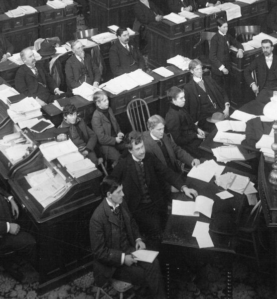 Detail of the Assembly Chamber photograph focusing on the journalists seated near the front of the room. During the 19th century, coverage of Capitol news received more attention from the state's newspapers than today. Of special interest is the female reporter in the center of the group and the three young pages. Architectural details revealed by the close view include the carpet pattern, the lamp on the clerk's table, and the Jenny Lind style tables at which the reporters are sitting.