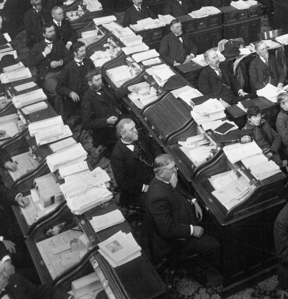 Detail of the 1899 Assembly Chamber photograph, showing a row of legislators at their desks. Until well into the 20th century, individual legislators had neither offices nor staff, and the result was the cluttered condition seen here. The roll top desk is closed for one absent member allowing a glimpse of the original hardware.