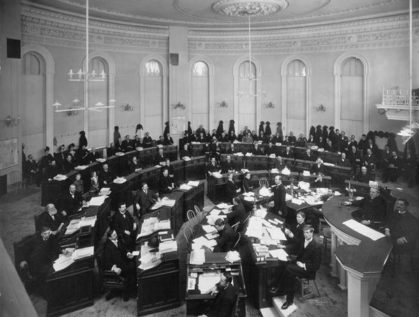 The only known visual documentation of the Senate Chamber in the third Wisconsin Capitol. It was probably more colorful than it appears here. In 1869 a reporter described the Senate walls as stippled in delicate lilac, with a white ceiling, and cornice ornamentation in pink. Maude Keene Gill, the daughter of an assemblyman, who saw the Senate during the early 1880s, remembered only that the room was done in white and pink. She was more impressed with the large gas chandeliers. "I hope I kept my mouth closed the first time I saw the man light it; it was a work of time, with a contrivance which first turned on the gas, and then lit it with a taper." The black & white 1899 photograph does not answer the question of whether those descriptions matched the circumstances in 1899.