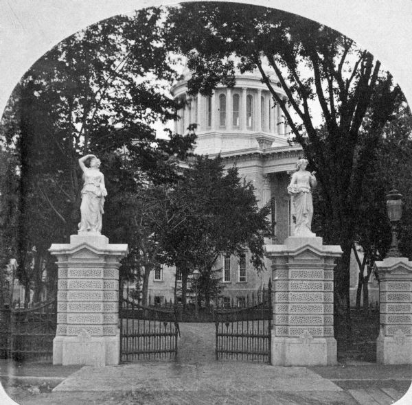 The southeast entrance to the Capitol Park at Monona Avenue (now Martin Luther King Boulevard), probably taken about 1875. In the foreground are elaborate stone gateways topped with statues which flank the sidewalk and iron gates. In the background is the Wisconsin State Capitol. This photograph was part of the "Beauties of the City of Lakes" stereograph series distributed by E.R. Curtiss.
