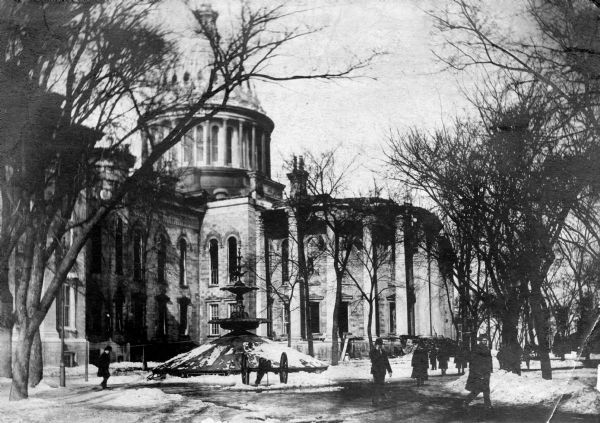 Damage caused to the East Wing of the third Wisconsin State Capitol by the fire of February 26-27, 1904. In the foreground is a duplicate of the "Centennial Fountain," which had won a medal at the U.S. Centennial Exposition in Philadelphia, in its wintertime covering. The fountain was moved to the governor's residence on East Gilman Street in 1912 and in 1943 the state sold the fountain for scrap to support the war effort.