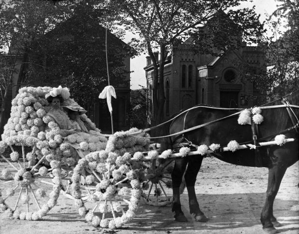 A horse-drawn carriage decorated for a floral parade on West Washington Avenue. The carriage is passing in front of the Gates of Heaven Synagogue (the building to the right).