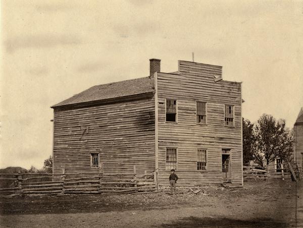 Council House of the first Wisconsin Territorial Legislature when it met at Belmont in 1836. A young boy or man wearing a hat stands in front near a fence.