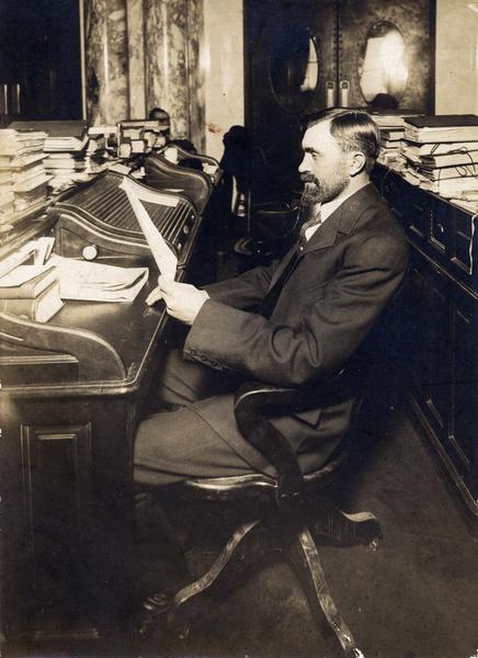 Otto Onstad, a newly elected assemblyman from Dane County, in the newly constructed Assembly Chamber of the present Wisconsin State Capitol. He is sitting at a row of desks from the third Wisconsin State Capitol, suggesting that a substantial number were rescued from the flames during the fire of 1904. The old desks were sold in 1913 when new furniture was purchased. However, two old desks are on display in the Assembly hall.