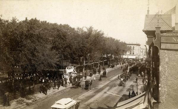 Military parade on Main Street honoring President Grover Cleveland's visit to Madison. Soldiers are marching to meet the President and crowds are watching from the Capitol Park which is decorated for the occasion.