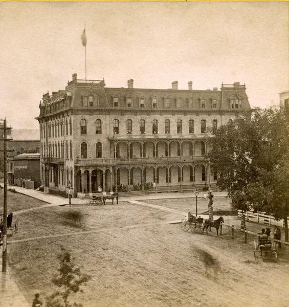 Elevated view of the Park Hotel at the intersection of Carroll and Main streets, with the Capitol Park on the right. Several carriages are hitched at the park railing. Because the wooden Capitol fence was replaced in 1873, this picture must have been taken no later than that year.