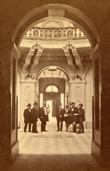 View of the only known view of the first floor Rotunda of the third Wisconsin State Capitol, a remarkable stereograph by Dane County photographer Andreas Larsen Dahl. A group of seven men in suits and hats stand together, and there appears to be a dog sitting at one man's feet. Details documented here include the white marble and black slate floor tiles, the enormous brackets that supported the second floor gallery, the gallery balustrade, and the Italianate detailing of the pillars. Careful observers can look through the Rotunda and see a chandelier and a ceiling medallion that match one in Image ID: 23110 which was photographed after the 1904 fire.