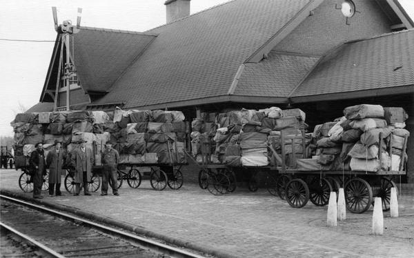 View across railroad tracks of cart loads of mail sacks on the platform, awaiting the arrival of the next train. The four men standing on the left have been identified as B. Black, L. Frazier, Wm. McLaughlin, and G. Schaad.