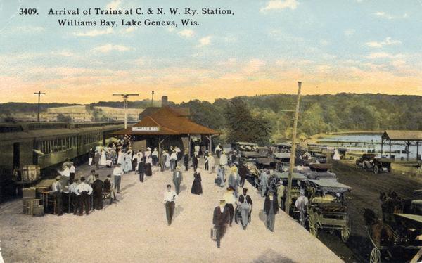 Color postcard of elevated view of passengers arriving on the Chicago & Northwestern Railroad train at the Williams Bay station. Many automobiles are parked near the depot, and the lake is in the distance.