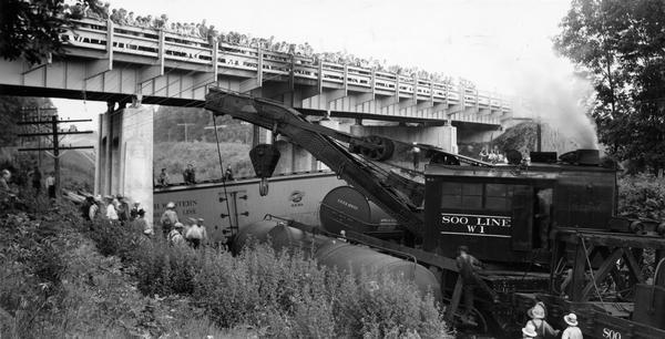 Derailment of a Minneapolis, St. Paul and Sault St. Marie Railway train near Waupaca. A large crowd is gathered on an overpass to watch the Soo Line's Wrecker W-1 lift the cars back on the track.
