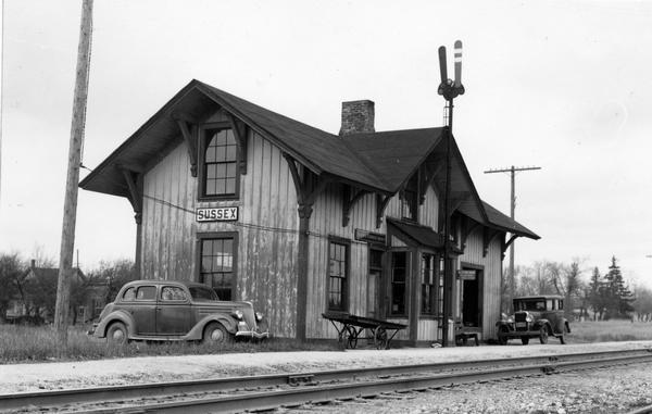 View across railroad tracks of the Wisconsin Central Railroad depot at Sussex (formerly known as Templeton). Two cars are parked near the depot and a baggage cart.