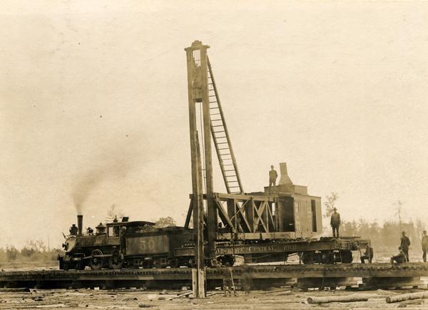 Wisconsin Central locomotive No #50 and pile driver No #2 working near Owen. Men stand on and around the tracks.