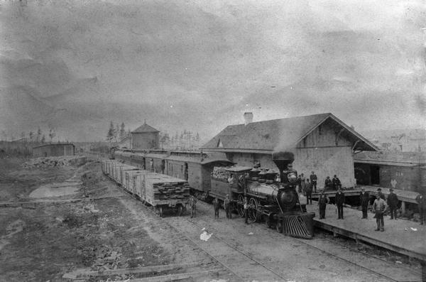 Elevated view of the Wisconsin Central Railroad passenger depot at Stuntz Avenue. A group of people stand on the platform and near the locomotive between the double set of railroad tracks. The locomotive pulling the train is No #3 which was built by the Baldwin Locomotive Workers and purchased by Wisconsin Central about 1871. The roundhouse is on the left in the background, with the wood supply for the engines in front of it.