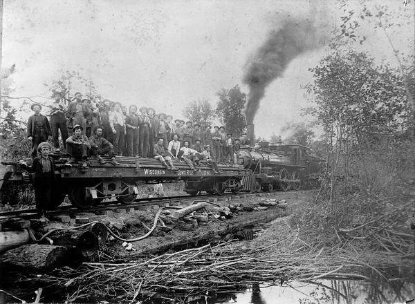 A work crew of the Upham Lumber and Manufacturing Company near Athens, Wisconsin. The men are riding on a flatbed car of the Abbottsford and Northeastern Railroad, a subsidiary of the Wisconsin Central Railway, for whom they will be constructing a new logging spur. The engine, (No #1000) was built by the Baldwin Locomotive Works and originally identified as No #28 of the Wisconsin Central Railway. The engine was sold to Upham in 1898 and then sold by Upham to the United States Leather Company in 1908 for use in the Rib Lake area. The draw bar attachments to flat car No 581 were patented by Williamson and Price in 1897. Railroad historian Roy L. Martin, from whose collection this image comes, noted the typical corduroy roadway and makeshift log bridge.