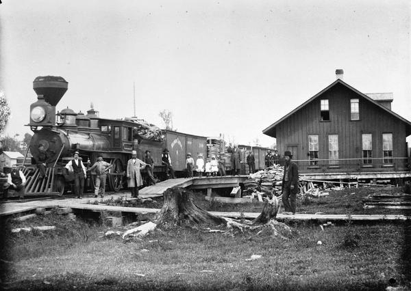 Group of men posing in front of the Wisconsin Central Railroad train at a depot.

Railroad historian Roy L. Martin, from whose collection this image comes, notes that locomotive No #6 (known as the Portage) was built by the Baldwin Locomotive Works in 1872. No 6 was one of the Baldwin engines of the Wisconsin Central. This locomotive was referred to as a "cold water Baldwin" because of a mechanical water pump which forced cold water into the boiler. Here the engine retains the crosshead boiler feed pumps. The engine also had brass bands around the steam chests and boiler jackets, broad bands around the front and back domes, and gold striping on the center dome and tender. The mudguards over the driving wheels were referred to as "pants."

Station agent Alex Mohr is near the door of the depot. He and his family lived above the depot, as was then the custom. The man in the linen duster is thought to be Gavin Campbell, superintendent of the road. The conductor is Tom Clark, the engineer is George Holmes.