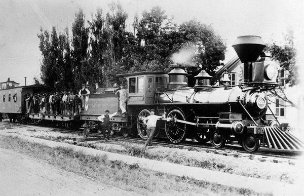 "The old wood train." It is a class H3 train built in 1880 by Manchester Locomotive Works. John Rhine, the engineer, is shown. A large group of men are standing on an open rail car behind the locomotive.