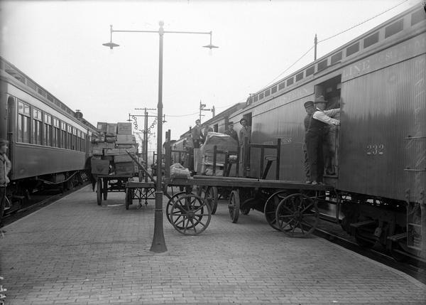 View down platform between two railroad trains of men unloading freight onto wagons from a Chicago, St. Paul, Minneapolis & Omaha Railway freight car at the Madison depot.
