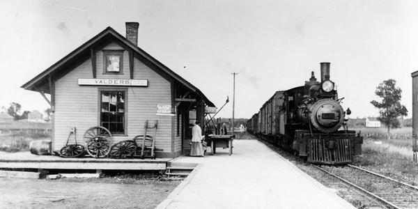 A single passenger awaits the arrival of a train at the Valders station. The train, which is a mixed local on the Manitowoc line, is being pulled by Minneapolis, St. Paul and Sault Ste. Marie Railway locomotive #2120, pulling a mixed local train on the Manitowoc Branch.  This engine, a Mogul 2-6-0, was built by the the Baldwin Locomotive Works in 1886 and scrapped in 1927.