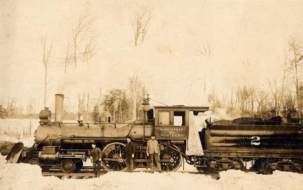 Three men stand in the snow in front of the Wisconsin & Northern Railroad engine #2 (the second locomotive with this number) in the woods on the Menominee Indian Reservation. The line, 125 miles long from Neenah to Argonne, was built by lumber and wooden ware interests in Neenah and Menasha, in order to harvest the timber between these points. Engine #2 was purchased from the Chicago, Milwaukee, St. Paul & Pacific Railway.