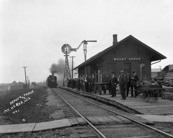 View across railroad tracks of passengers standing on the platform as a locomotive pulls into the depot.