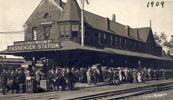View across railroad tracks of the original Chicago and North Western Railroad passenger station, 219 South Blair Street at East Wilson Street. A large crowd of people are waiting on the platform. Torn down in 1910.