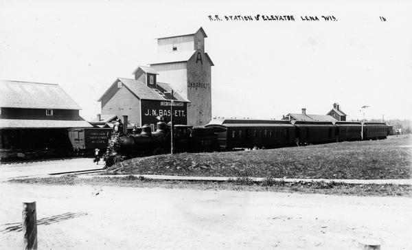 The Chicago, Milwaukee, & St. Paul Railroad depot and the J.N. Bassett grain elevator. A four-car passenger train is stopped at the station.