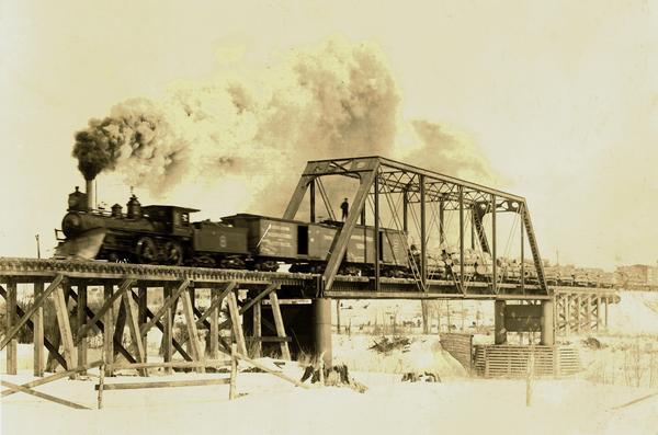 View from shoreline of the Fairchild & Northeastern steam locomotive pulling a load of logs over the Black River bridge near Greenwood. A man is standing on top of one of the boxcars.