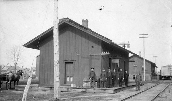 View across railroad tracks towards the railroad depot. The employees posed in front of the depot are identified only by their last names, left to right: Laabs, warehouseman; Ferguson, warehouseman; Goodrich, agent; Tuttle, operator; Haas, baggage man, Brown, bill clerk; and an unidentified car repairman.