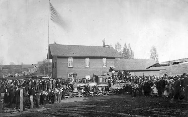 The arrival of the first train in Waukon, on the Wisconsin & Michigan Railroad line. There is a large crowd gathered near the tracks, and one man stands on the roof of the railroad depot.