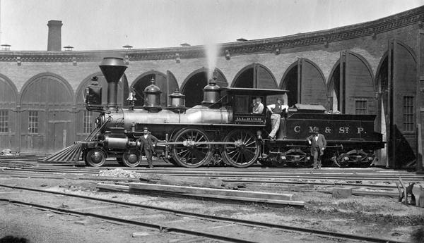 The locomotive "D.K. Bush," which was built in the Racine shops of the Western Union Railroad in 1879, the same year in which the line came into the possession of the Chicago, Milwaukee & St. Paul Railroad. On the ground, against the tender, is master mechanic John Taylor, in the cab doorway is Mart Gorman and, leaning against the connecting rod is D.L. Bush himself, then assistant superintendent of the southwestern division.