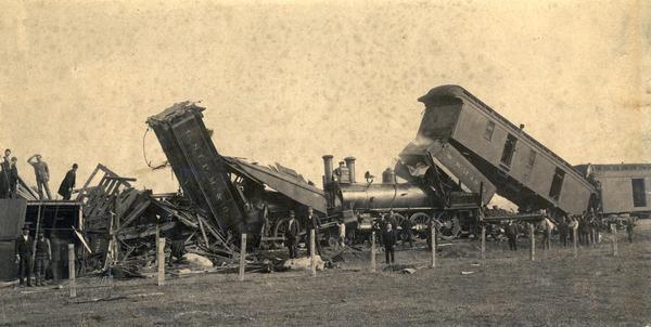 The wreckage of a head-on collision of two Chicago, St. Paul Minneapolis & Omaha Railroad trains at Tramway. Men stand near and on top of the railroad cars. In the foreground is a field and fence, and dead cows are lying near the railroad cars.
