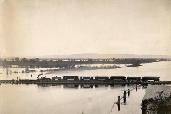 Elevated view of the pile-pontoon railroad bridge across the Mississippi River at Prairie du Chien. The bridge was built and patented by John Lawler in 1874 and it solved the problem of providing railroad crossing of the Mississippi without stopping the river traffic. Prior to the Lawler bridge, the railroad cars were towed across the river on barges. The Lawler Bridge was 8000 feet long, crossing both channels of the river and an intervening island.  It was constructed in two parts, the pile, or stationary part, and the pontoon or movable part, which consisted of two floating draws, one in each channel, which, when closed, form an unbroken track, permitting a safe and rapid railroad crossing. When open, the bridge allowed river traffic to pass with ease. The pile portion of the bridge was of ordinary railroad construction, but the draw over each channel consisted of one pontoon with a 12 inch draft. The Lawler bridge was replaced in 1910.